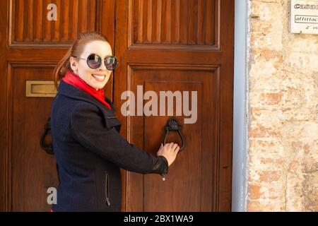 Une femme souriante en lunettes de soleil, une veste noire, une écharpe rouge et un pantalon vert frappe sur une vieille porte en bois d'époque d'une maison italienne. Élégant Banque D'Images