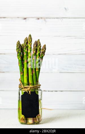 Asperges fraîches dans un pot en verre sur fond de bois blanc avec étiquette vierge en tableau noir Banque D'Images