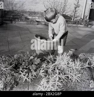 Années 1960, historique, à l'extérieur sur un jardin de patio, un jeune garçon avec une petite truelle jouant avec la terre dans un pot en terre cuite, Angleterre, Royaume-Uni. Banque D'Images