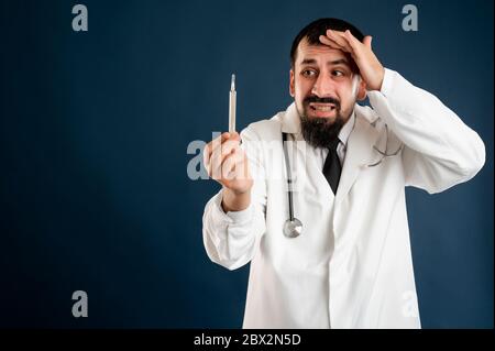 Portrait d'un médecin masculin avec stéthoscope en uniforme médical est en regardant le thermomètre posé sur un fond bleu isolé. Banque D'Images