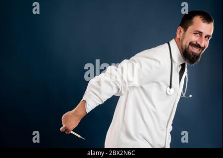 Portrait d'un médecin masculin avec stéthoscope en uniforme médical plaisantant avec thermomètre posé sur un fond bleu isolé. Banque D'Images