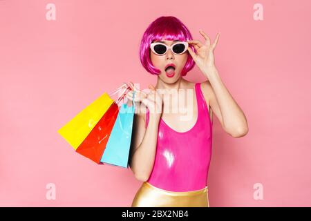 Image d'une fille surprise portant une perruque et des lunettes de soleil tenant des sacs de shopping isolés sur fond rose Banque D'Images