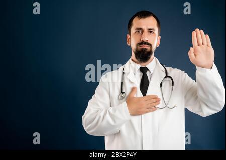 Portrait d'un médecin masculin avec stéthoscope en uniforme médical montrant le serment posé sur un fond bleu isolé. Banque D'Images