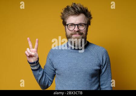 Positif homme dans les lunettes fait un V-signe avec les doigts. Boxer à la main, un hipster barbu dans un ancien pull fait un geste de victoire. Banque D'Images