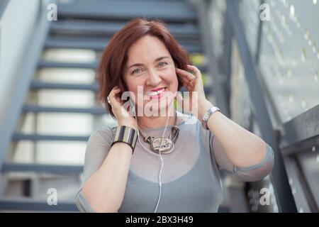 Femme élégante et excitée dans un casque d'écoute, qui écoute de la musique dans la rue en souriant, qui regarde la caméra, heureuse de porter un chemisier gris Banque D'Images
