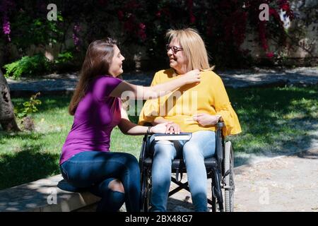 Mère en fauteuil roulant et sa fille tenant les mains et souriant les unes aux autres sur un fond défocayeux. Handicap et concept de famille. Banque D'Images
