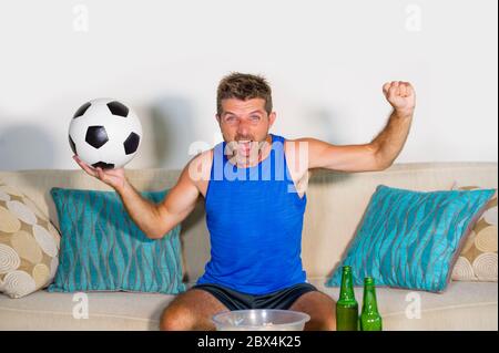 Jeune homme attrayant heureux et excité regardant un match de football à la télévision célébrant le but de victoire fou et spastique avec la bière pop-corn et le ballon de football dans f Banque D'Images