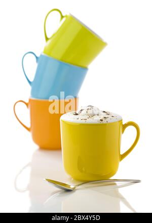 tasse jaune remplie de cappuccino, cuillère à l'avant, tasses vertes, orange et bleues empilées en arrière-plan, sur une surface réfléchissante blanche Banque D'Images