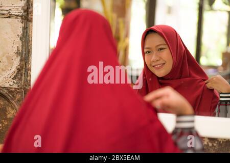 Miroir reflet de la jeune heureuse et belle musulmane dans hijab traditionnel ajustant son foulard rouge tête joyeuse poupées en beauté et de fashi Banque D'Images