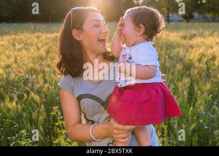 Une jeune mère tient sa petite fille dans les bras comme ils rient ensemble dehors un jour ensoleillé. Banque D'Images