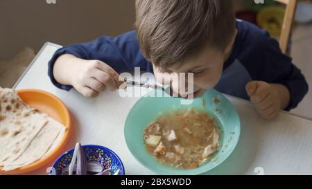 Un petit garçon d'âge préscolaire affamé mangeant de la soupe dans un bol vert. Un charmant enfant du Caucase se mange lui-même. Vue grand angle Banque D'Images