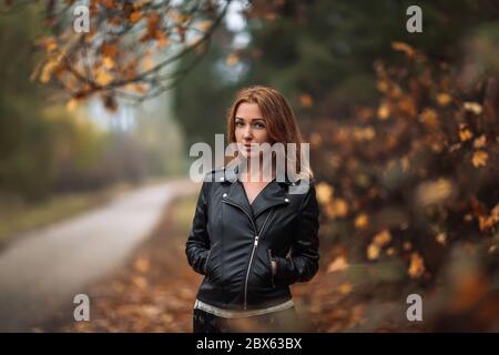 Femme rêveuse à tête rouge avec de longs cheveux bouclés sur un arrière-plan automnal flou. Fille sur fond fabuleux de parc avec des feuilles d'automne orange et roa Banque D'Images