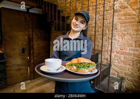 Gros plan sur une belle serveuse caucasienne souriant, tenant avec joie un plateau avec une tasse de délicieux café chaud et fraîchement cuit Banque D'Images