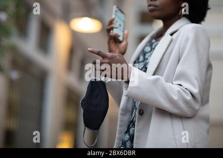 Un masque facial en tissu et un téléphone portable dans la main d'une personne noire. Les masques faciaux sont utilisés pour arrêter la propagation du coronavirus COVID-19 pandémique Banque D'Images