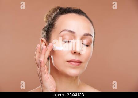 Jolie femme aux yeux fermés appliquant de la crème sur la zone sous-oculaire de son visage tout en prenant soin de la peau après l'hygiène du matin en isolement Banque D'Images