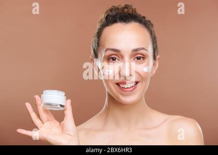 Bonne jeune femme magnifique avec un sourire éclatant tenant un petit pot de crème hydratante et rajeunissante devant l'appareil photo Banque D'Images