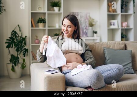 Bonne jeune femme enceinte à pattes croisées qui regarde une petite chemise pour un futur bébé dans ses mains tout en étant assise sur un canapé dans le salon Banque D'Images