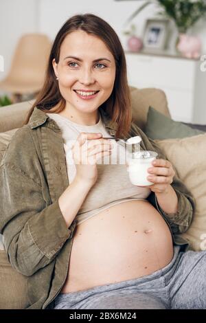 Jeune femme enceinte souriante en tenue décontractée, mangeant du yaourt tout en se relaxant sur le canapé et en appréciant du temps libre à la maison Banque D'Images