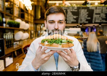 Faites une pause, déjeuner. Gros plan sur un joli jeune barbu, tenant une assiette avec un croissant frais et en appréciant sa pause tout en sentant Banque D'Images