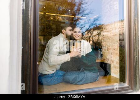 Un couple heureux prenant le petit déjeuner assis près de la fenêtre dans le confortable café de la ville, boire du café et manger des croissants traditionnels délicieux. Vue à travers Banque D'Images