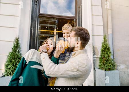 Famille heureuse, père, mère et enfant mignon, debout ensemble et s'amuser à l'extérieur, en appréciant leur déjeuner et en mangeant des croissants frais Banque D'Images