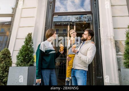 Famille heureuse, père, mère et enfant mignon, debout ensemble et s'amuser à l'extérieur, en appréciant leur déjeuner et en mangeant des croissants frais Banque D'Images