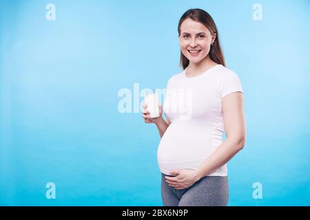 Bonne jeune femme avec un verre de lait touchant son ventre enceinte tout en disant que le calcium est nécessaire pour la future mère et son bébé Banque D'Images