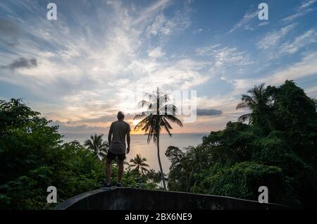 Silhouette d'homme debout seul sur la colline avec un cocotier et de palmiers et jouit de la mer sur un horizon dans un coucher de soleil. Concept de vacances et de voyages sur un Banque D'Images