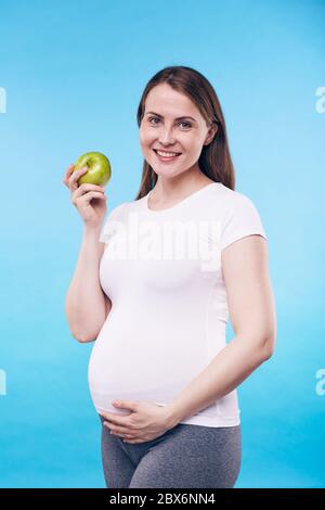 Bonne femme enceinte avec une pomme granny smith touchant le bas de son ventre tout en se tenant devant l'appareil photo en isolement Banque D'Images