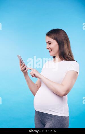 Bonne jeune femme enceinte en appuyant sur le bouton sur l'écran du smartphone tout en faisant selfie ou en téléphonant quelqu'un en isolement Banque D'Images