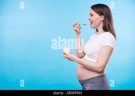 Jeune femme enceinte active avec une petite tasse en plastique de produit laitier appréciant son goût dans l'isolement avec l'espace de copie pour votre message Banque D'Images