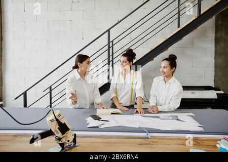 Groupe de trois jeunes créateurs de mode en chemises blanches remue-méninges et discutant des idées pour la nouvelle collection saisonnière lors de la réunion Banque D'Images