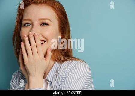 Belle fille à cheveux rouges dans une chemise rayée se tient sur un fond bleu et rit, se cachant derrière sa main. Banque D'Images