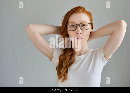 Une fille aux cheveux rouges dans un T-shirt blanc et des lunettes se tient avec les mains derrière la tête et levant un sourcil, regarde dans le cadre Banque D'Images