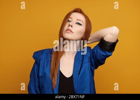 Une belle fille aux cheveux rouges avec de longues flèches et une veste bleue surdimensionnée se tient sur un fond jaune avec la main derrière la tête Banque D'Images