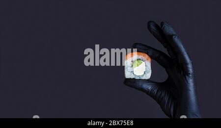 Concept alimentaire. Main femelle dans des gants en caoutchouc tenant le rouleau de saumon sushi sur un fond sombre avec copyspace. Menu de sushis. Cuisine japonaise. Banque D'Images