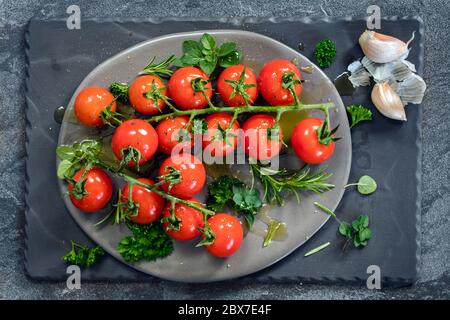 Tomates cerises sur la vigne avec des herbes fraîches, de l'huile d'olive, des épices et de l'ail, prêtes à cuire. Vue de dessus, sur ardoise. Banque D'Images