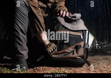 Homme en tenue tactique tenant un couteau et agenouillant pour sac à dos avec camping et équipement tactique sur fond de forêt nocturne. Banque D'Images