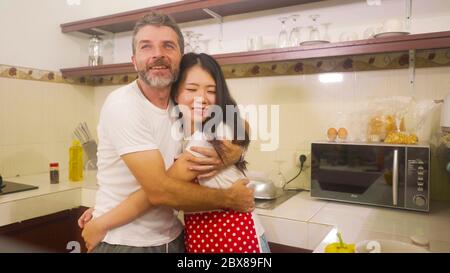 Jeune beau et heureux couple mixte d'origine ethnique dans l'amour cuisiner ensemble à la cuisine de la femme asiatique coréen en tablier rouge et son mari embrassant Banque D'Images