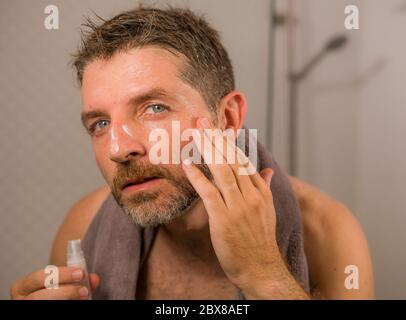 accueil style de vie portrait de jeune heureux et attrayant homme appliquant crème hydratante masque de visage traitement regardant le miroir de salle de bains dans l'homme anti vieillissement Banque D'Images