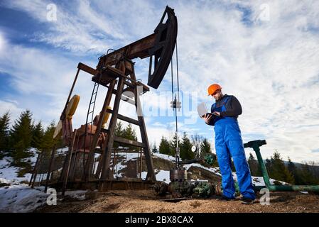 Homme utilisant un ordinateur portable pour contrôler le travail du cric de la pompe à pétrole. Combinaison de travail et casque de l'opérateur à proximité de l'unité de pompage d'huile. Concept de l'industrie pétrolière et de l'extraction de pétrole. Banque D'Images