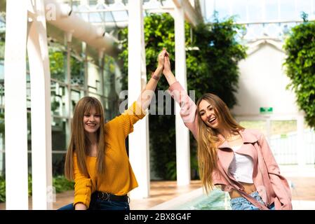 Deux jeunes filles caucasiennes attrayantes avec des cheveux blonds se secouer les mains avec leurs bras dans l'air en souriant à une fontaine à l'intérieur d'une pièce lumineuse avec le Banque D'Images