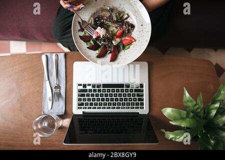 Photo en gros plan de la salade dans les mains et du verre avec couteau et fourchette sur la table au restaurant. Photo de salade et ordinateur portable sur table au café Banque D'Images