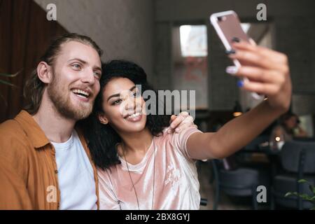 Jeune couple faisant des photos sur un appareil photo de téléphone portable frontal dans le restaurant. Belle femme afro-américaine faisant la photo avec un ami dans le café. Amis souriants hap Banque D'Images