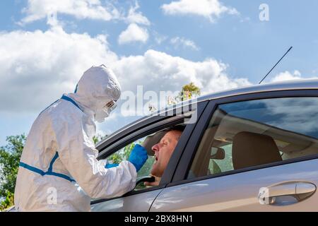 Un professionnel de la santé entièrement couvert d'un costume de protection blanc et de lunettes fait fonctionner un écouvillon pour Covid-19 à un homme assis à l'intérieur de sa voiture Banque D'Images