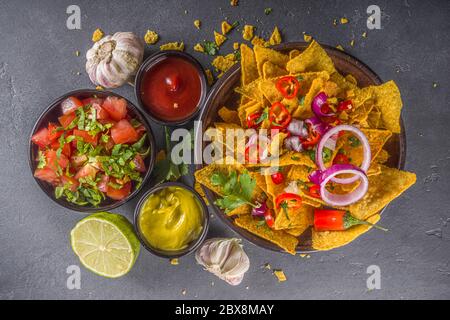 Croustilles nachos au fromage fondu et diverses sauces mexicaines traditionnelles, salsa et jalapeno, fond gris foncé vue du dessus espace copie Banque D'Images