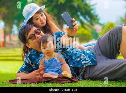 Jeune famille japonaise asiatique heureuse et aimante avec parents et petite fille douce au parc de la ville avec le père prenant le selfie pic avec téléphone portable Banque D'Images
