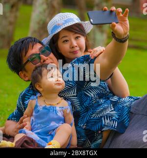 Jeune famille japonaise asiatique heureuse et aimante avec parents et petite fille douce au parc de la ville avec le père prenant le selfie pic avec téléphone portable Banque D'Images
