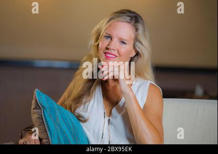 portrait de style de vie dans la faible lumière de belle et élégante femme blonde cheveux sur ses 40 s assis heureux à la maison salon détendu sur canapé canapé regardant ch Banque D'Images