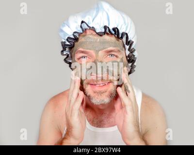 style de vie portrait drôle de l'homme étrange heureux sur la pomme de douche regardant à lui-même dans le miroir de salle de bains avec la crème verte sur son visage application masque facial peau Banque D'Images